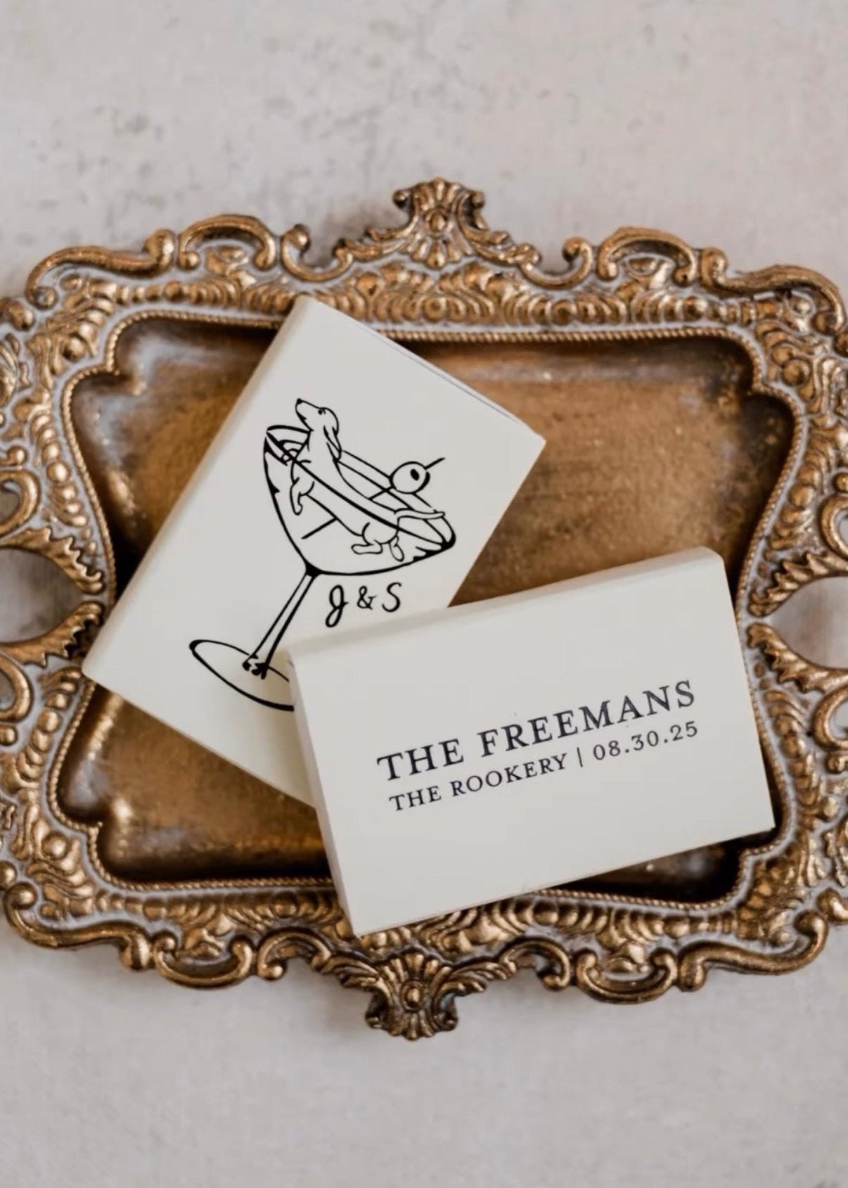 Two custom printed foil matchboxes cards on an ornate gold frame with a white background. Features a black and white illustration of a dachshund swimming in a martini glass with an olive pick, personalized with initials "J&S" and names, location, date "The Freemans, The Rookery, 08.30.25"